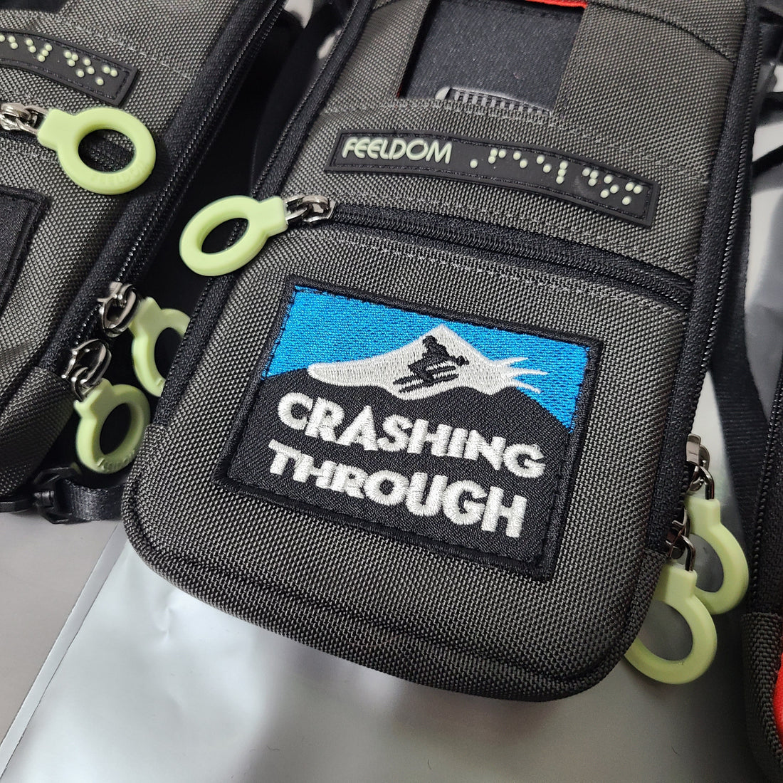 A gray phone pouch with a braille label and black, white and blue embroidered patch that says "crashing through" in bold letters, and a little skier on a mountain.