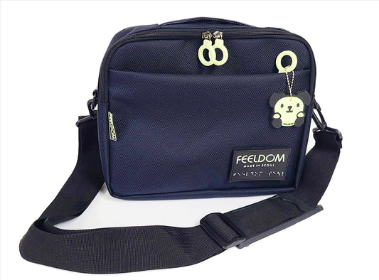 Dark Blue Rectangle pouch with double white zipper closure and front zipper pocket. A glow in the dark dog character rubber keychain and black rubber braille FEELDOM logo patch are on the front. THere is a wide black shoulder strap which is attached to the sides of the pouch. It is 11 inches by 8 inches tall, and 2 to 3 inches thick.