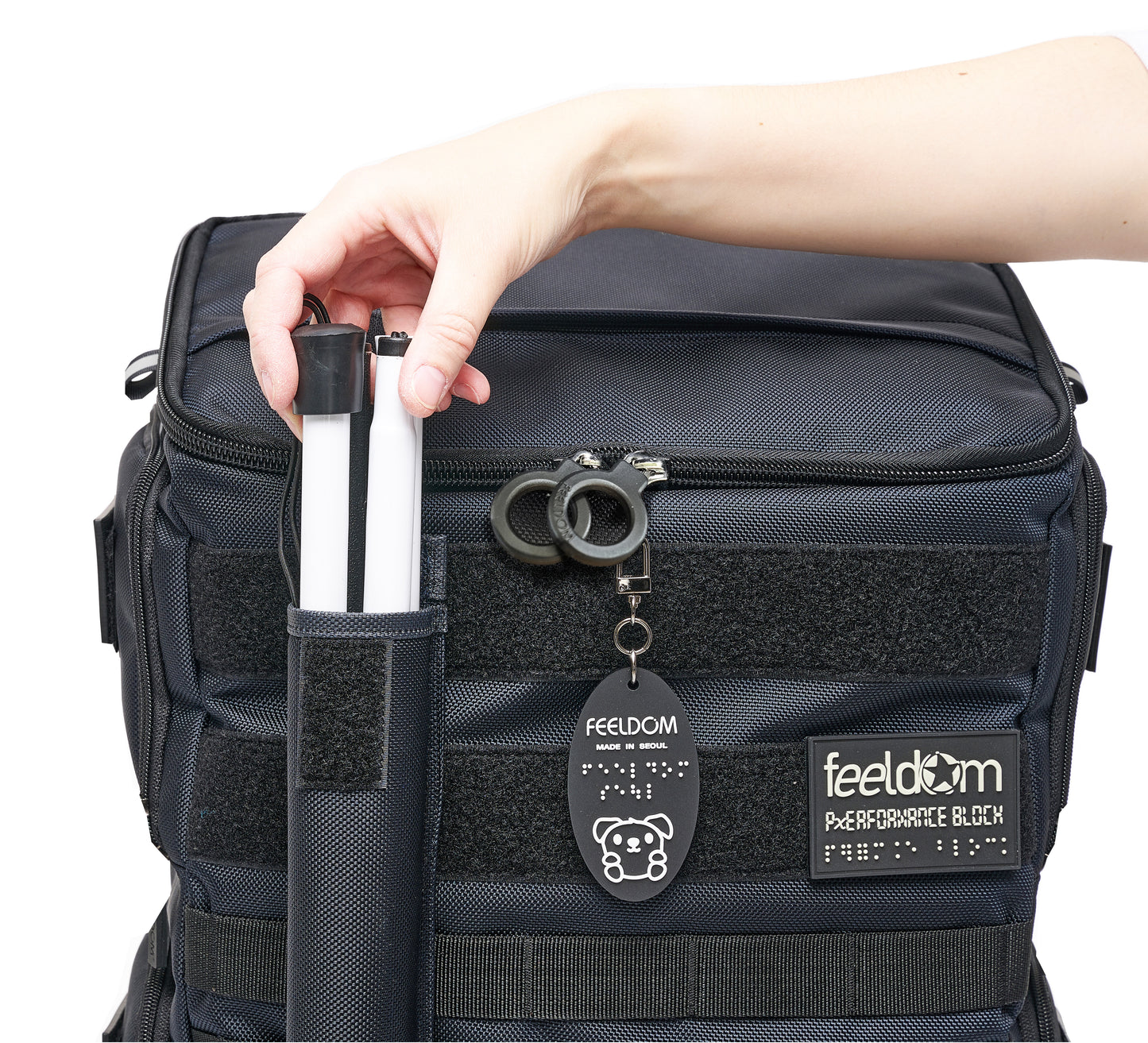 Performance Block has a nylon can pouch attached to the front with velcro. A hand reaches out to take out the white cane out of the pouch. The premium black plastic ring zippers say "Feeldom" on them, and the glow in the dark patch reads "Feeldom Performance Block" in Braille